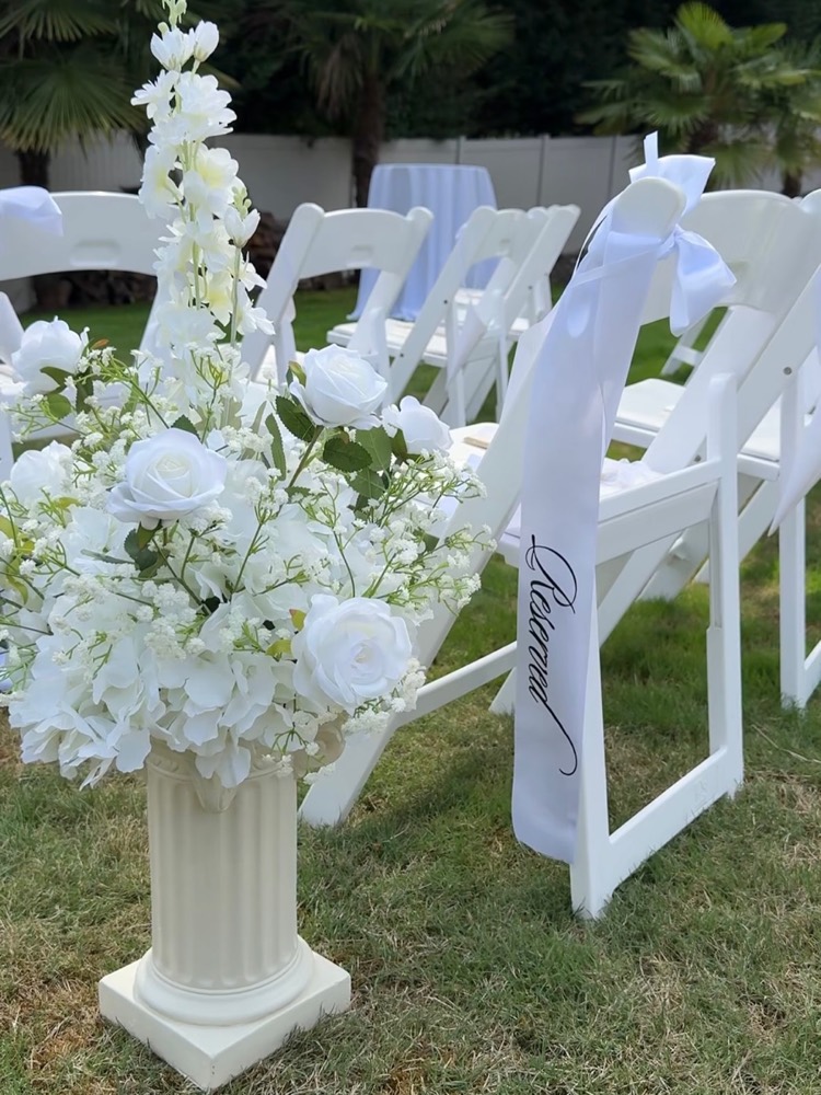 White floral aisle arrangement on a white pillar beside ceremony chairs